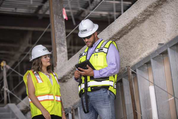 Women and man with white hard hats and safety vests looking at job progress on a tablet.