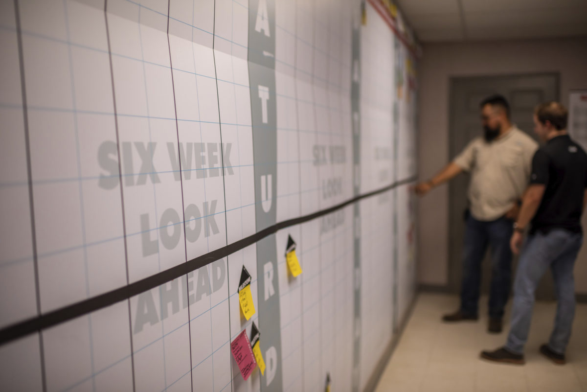 Two men looking at a large schedule and calendar with post-it notes on the wall.