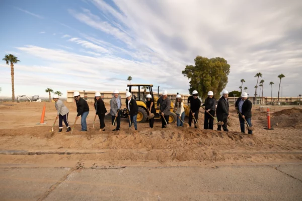 Twelve people, each wearing a white hard hat and holding a shovel, standing front of a bulldozer on a construction site.
