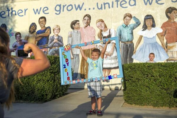 Young boy stand in front a mural of people for a first day of school photo while his brother looks over a hedge behind him.