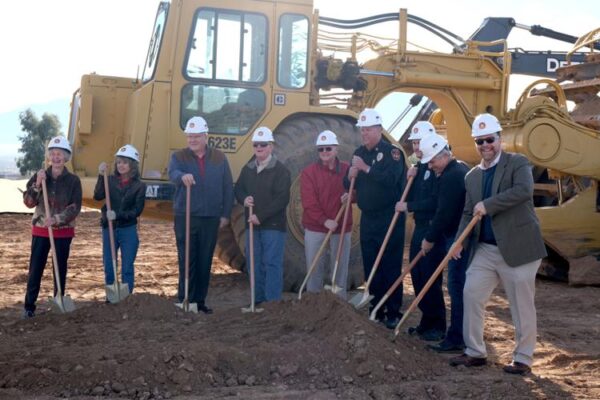 Nine men and women in white hard hats who are standing in front of a bulldozer and hold shovels on the construction site.