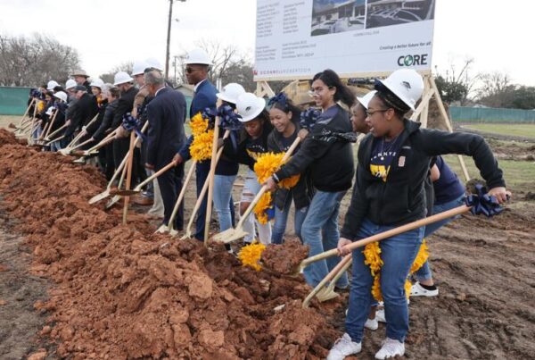 Middle school students and local officials with white hard hats hold shovels and stand on construction site dirt in a groundbreaking ceremony.