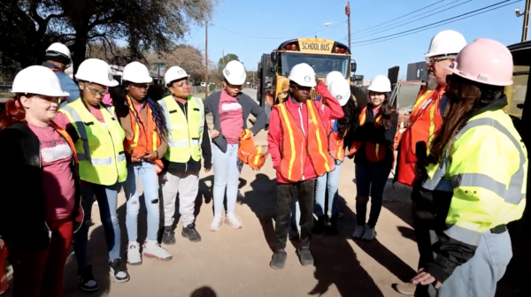 A group of students in white hard hats and safety vests prepare to tour the construction site.