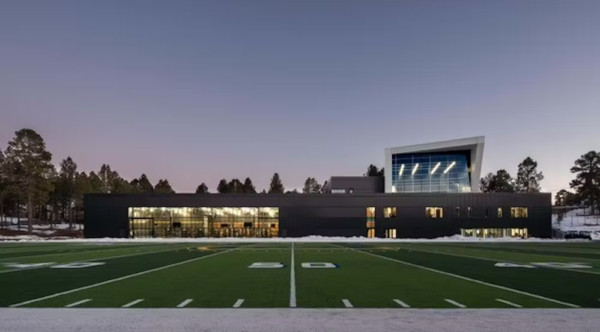A view of the practice football field at the 50-yard line at sunset, with the athletic facility in the background with its lights on.