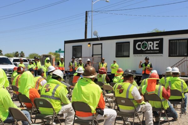 Large group of construction workers in hard hats and safety vests sitting in chairs during a safety outside the CORE Construction office on a construction site.